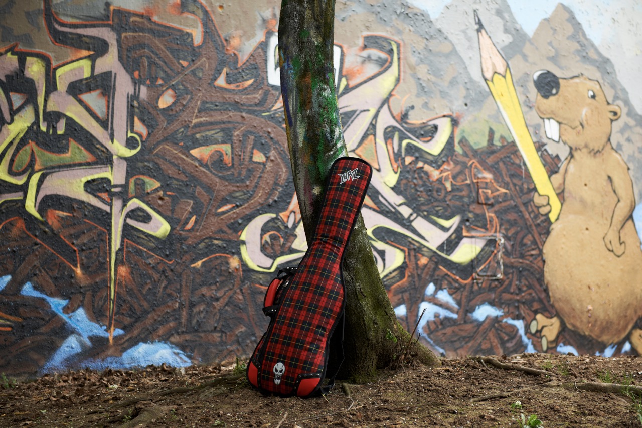 Red plaid guitar case with “Lørz” logo and skull emblem leaning against a tree trunk in front of a graffiti wall that includes a cartoon beaver holding a giant yellow pencil.