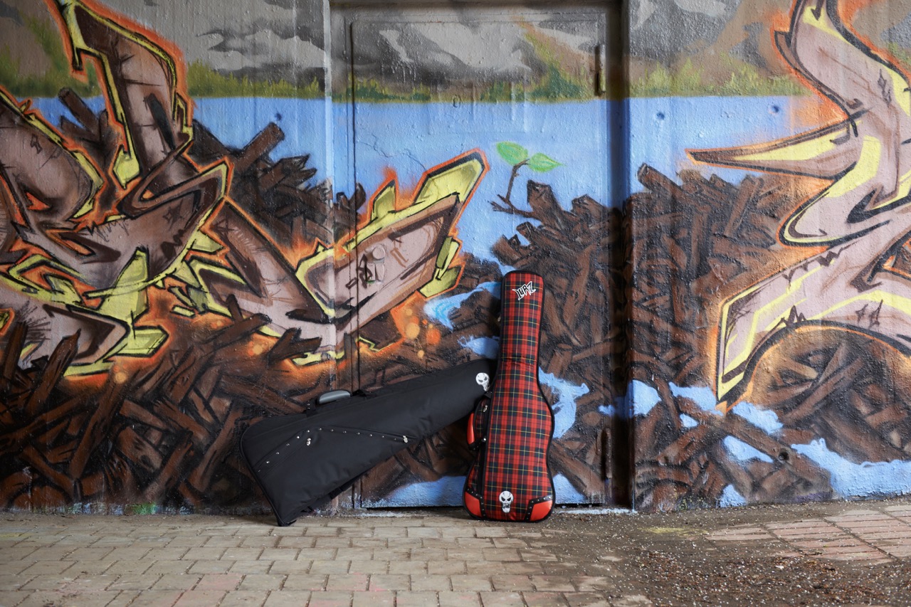 Red plaid guitar case with “Lørz” logo and skull emblem and a matching black guitar case leaning together against a vibrant graffiti wall featuring abstract art and a small green sprout.