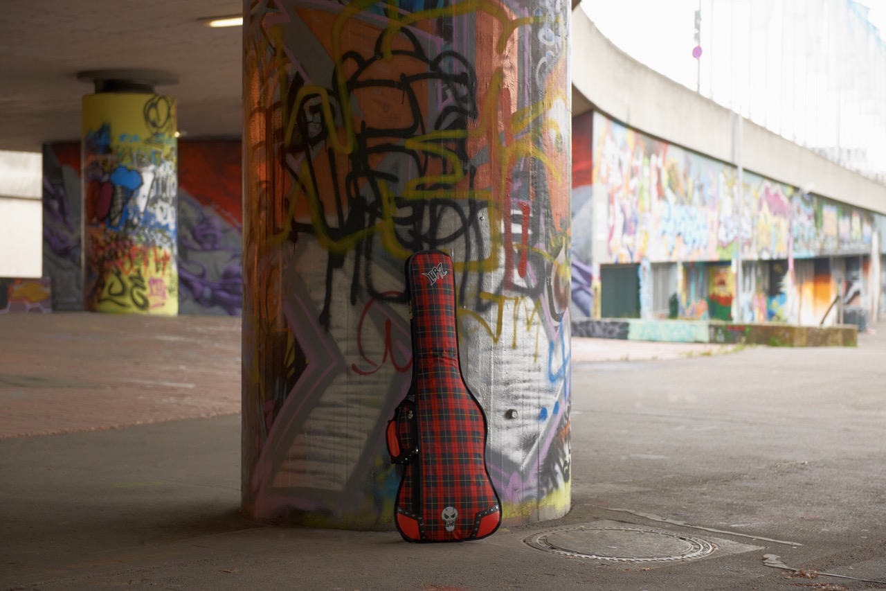 Red plaid guitar case with white “Lørz” logo and skull emblem leaning against a colorful graffiti-covered concrete pillar in an urban underpass or skatepark.