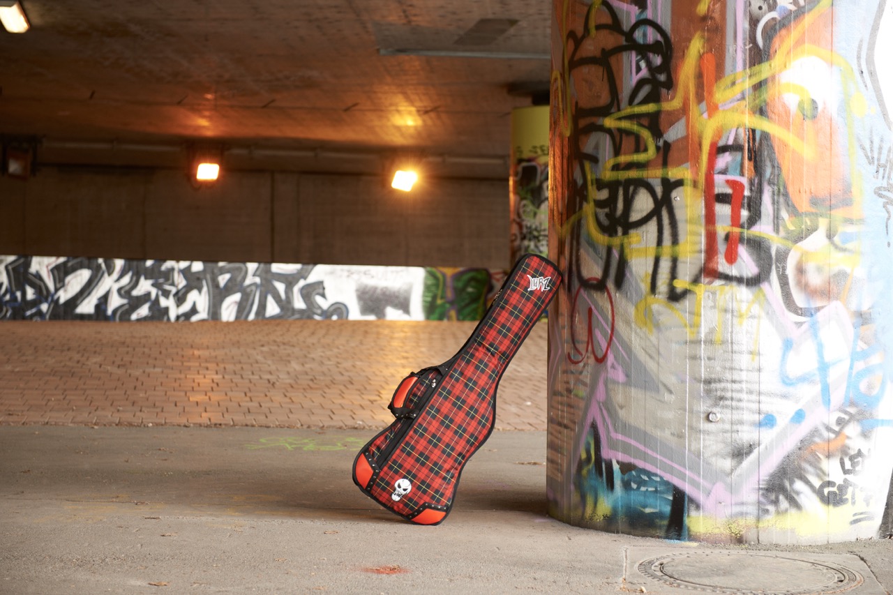 Red plaid guitar case with “Lørz” logo and skull leaning against a graffiti pillar inside a graffiti-covered urban underpass with brick flooring and warm overhead lighting.