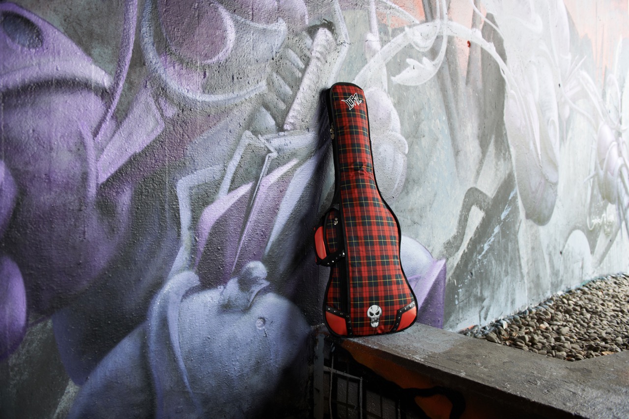 Red plaid guitar case with “Lørz” logo, skull emblem resting on a wooden ledge against a large purple-and-grey graffiti mural wall, with gravel ground beside it.