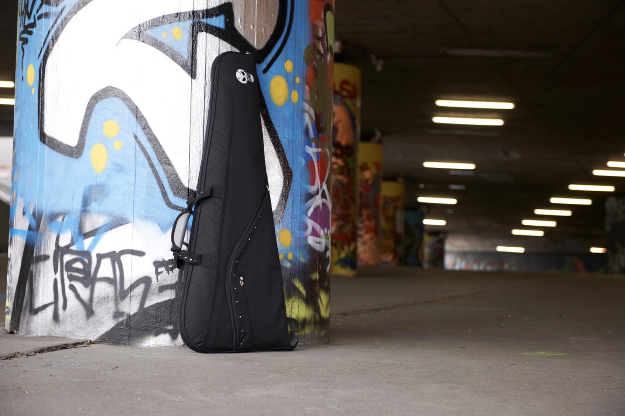 Black quilted guitar case with “Lørz” logo and skull emblem leaning against a large blue-and-white graffiti pillar inside a dimly lit concrete tunnel lined with fluorescent ceiling lights.